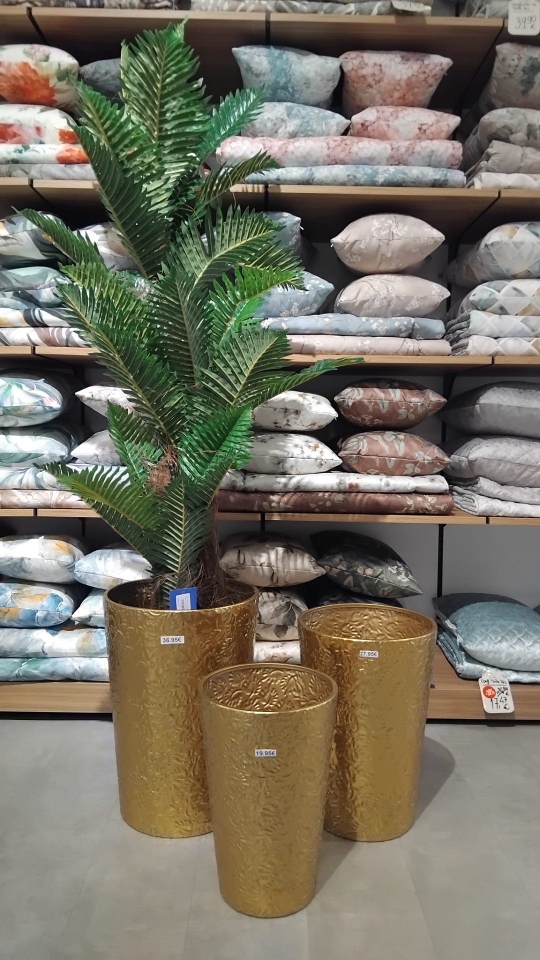 Três vasos dourados com planta artificial e prateleiras com almofadas e mantas