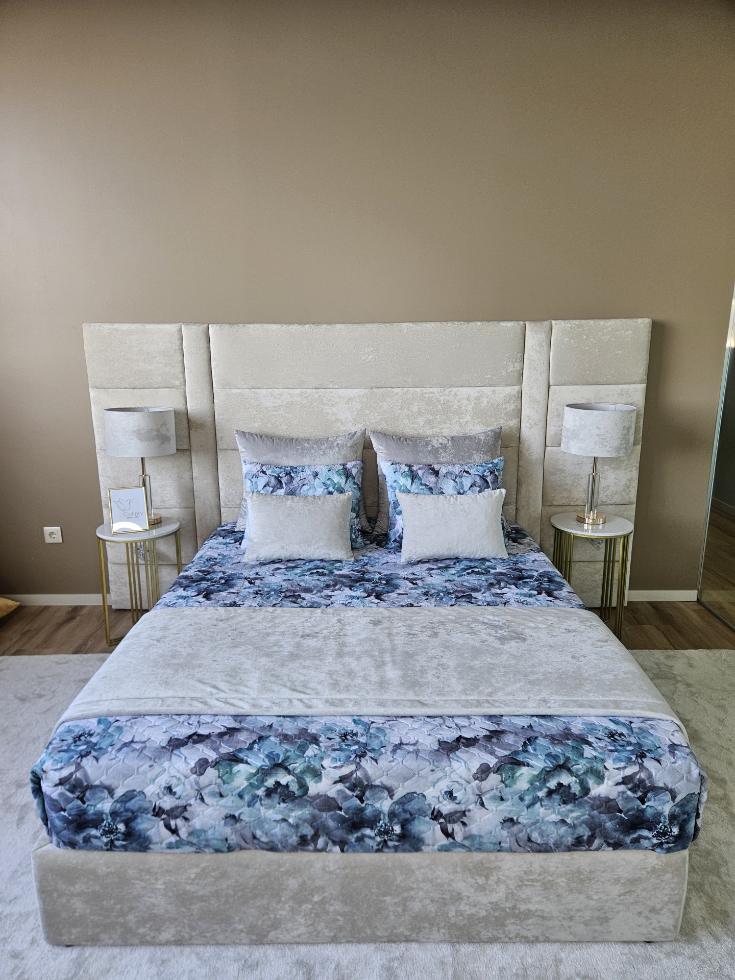 Cama com cabeceira cinza, edredão floral azul e cinzento com duas mesas de cabeceira douradas e lâmpadas cinzentas