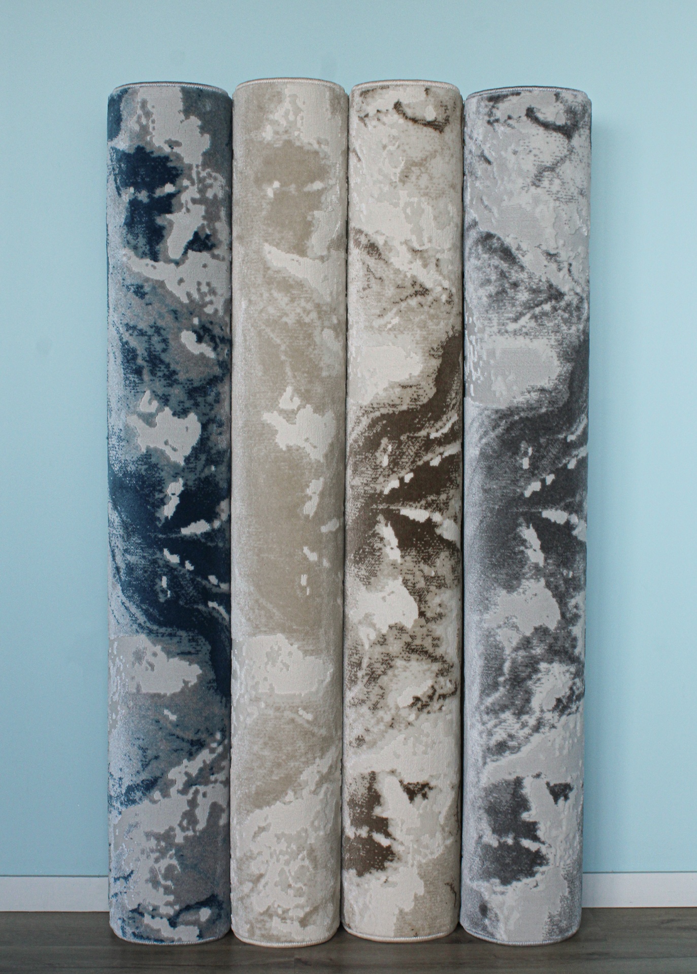 Tapetes enrolados com padrões abstratos em azul, bege, castanho e cinza encostados numa parede azul clara.