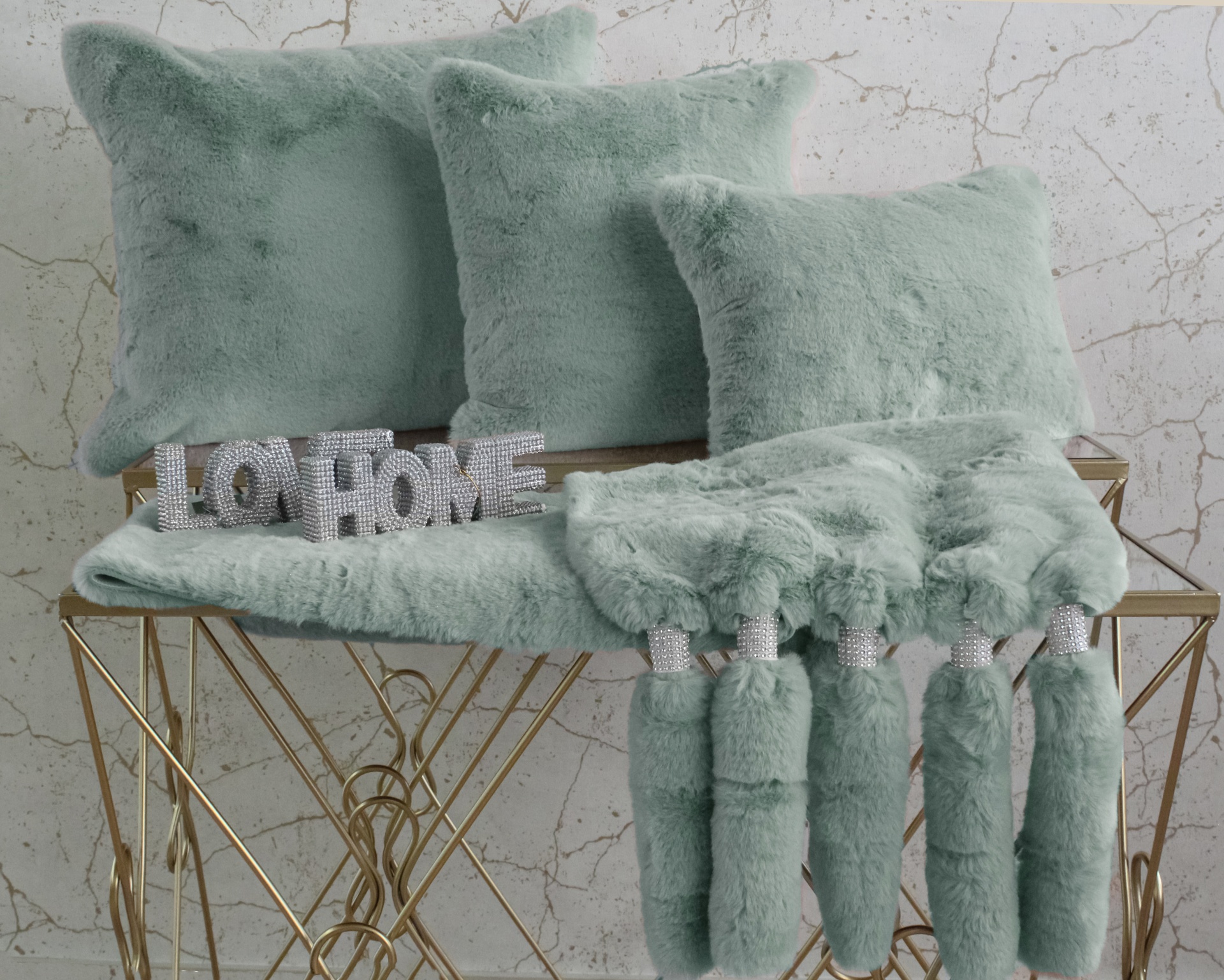 Três almofadas verdes de pelúcia e manta verde com franjas sobre mesa dourada com placa LO HOME