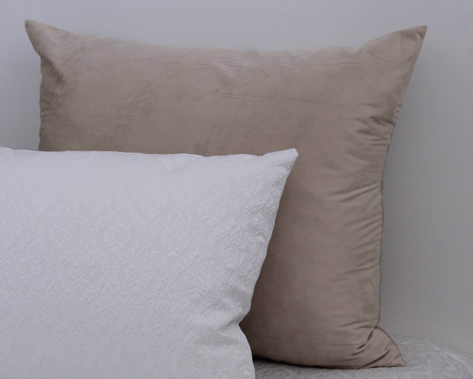 Duas almofadas, bege aveludada e branca com padrão em relevo, numa cama com roupa branca
