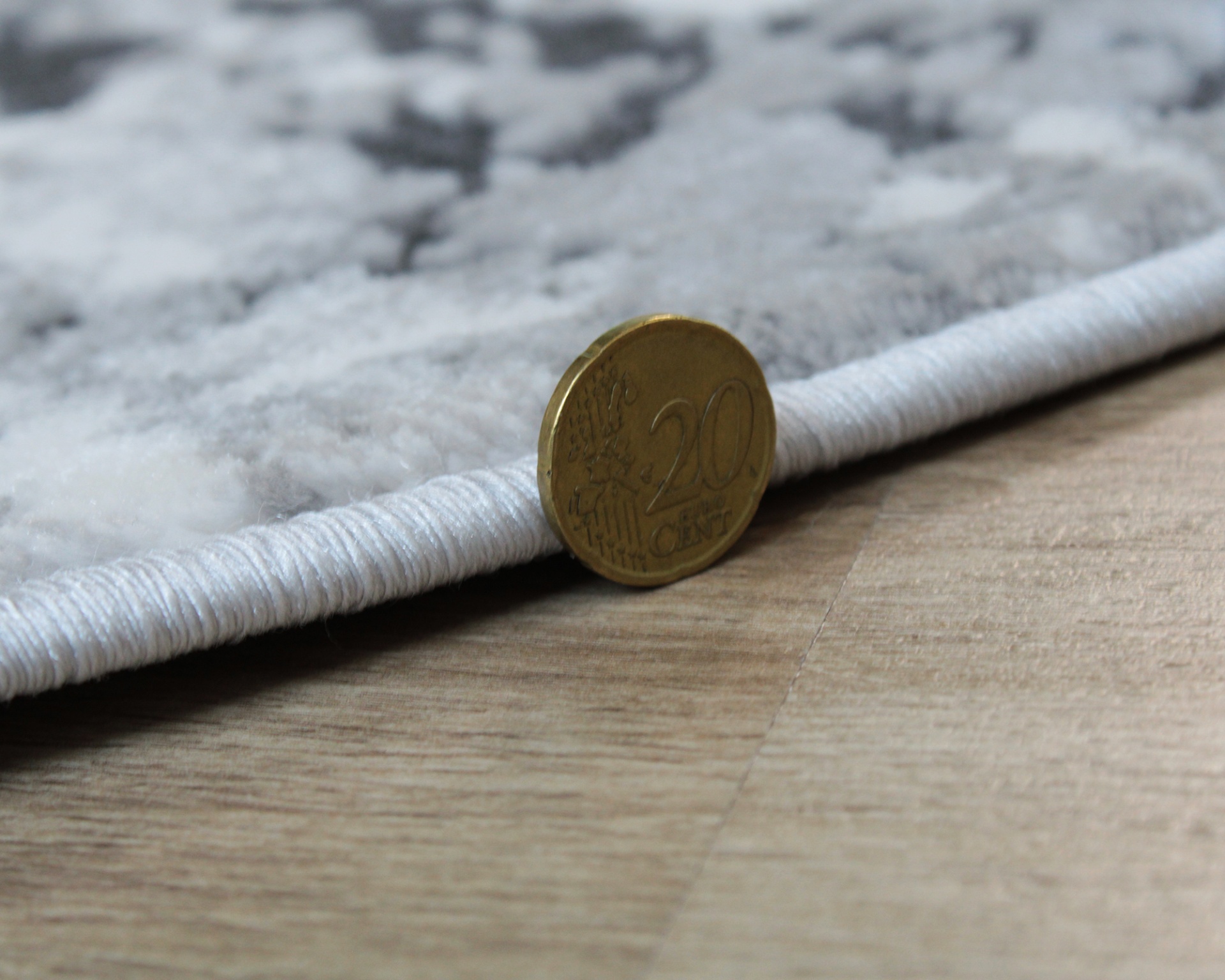 Moeda de 20 cêntimos ao lado da borda de tapete cinzento e chão de madeira.