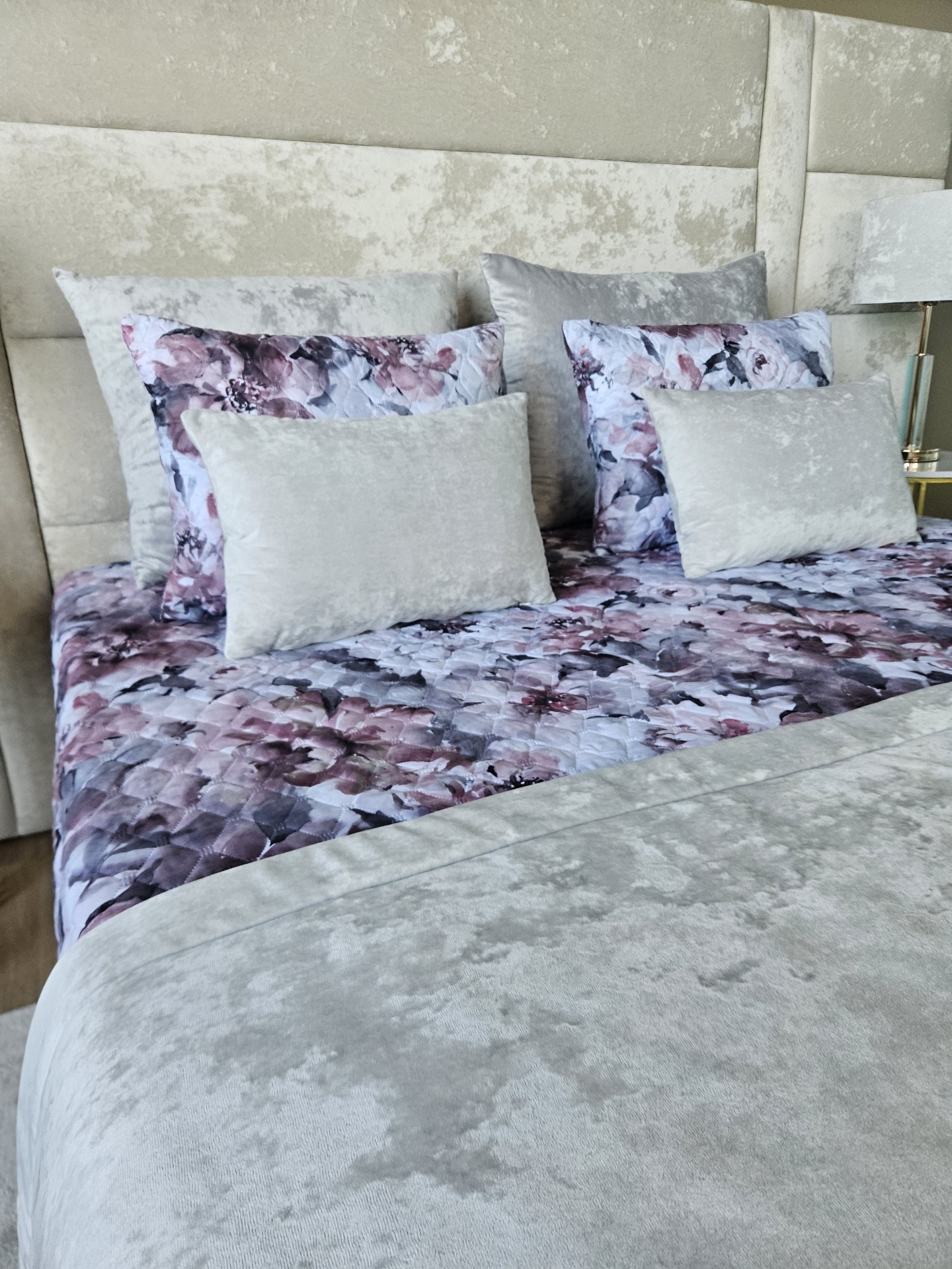 Conjunto cama com edredão floral roxo, almofadas cinzentas e cabeceira aveludada cinzenta
