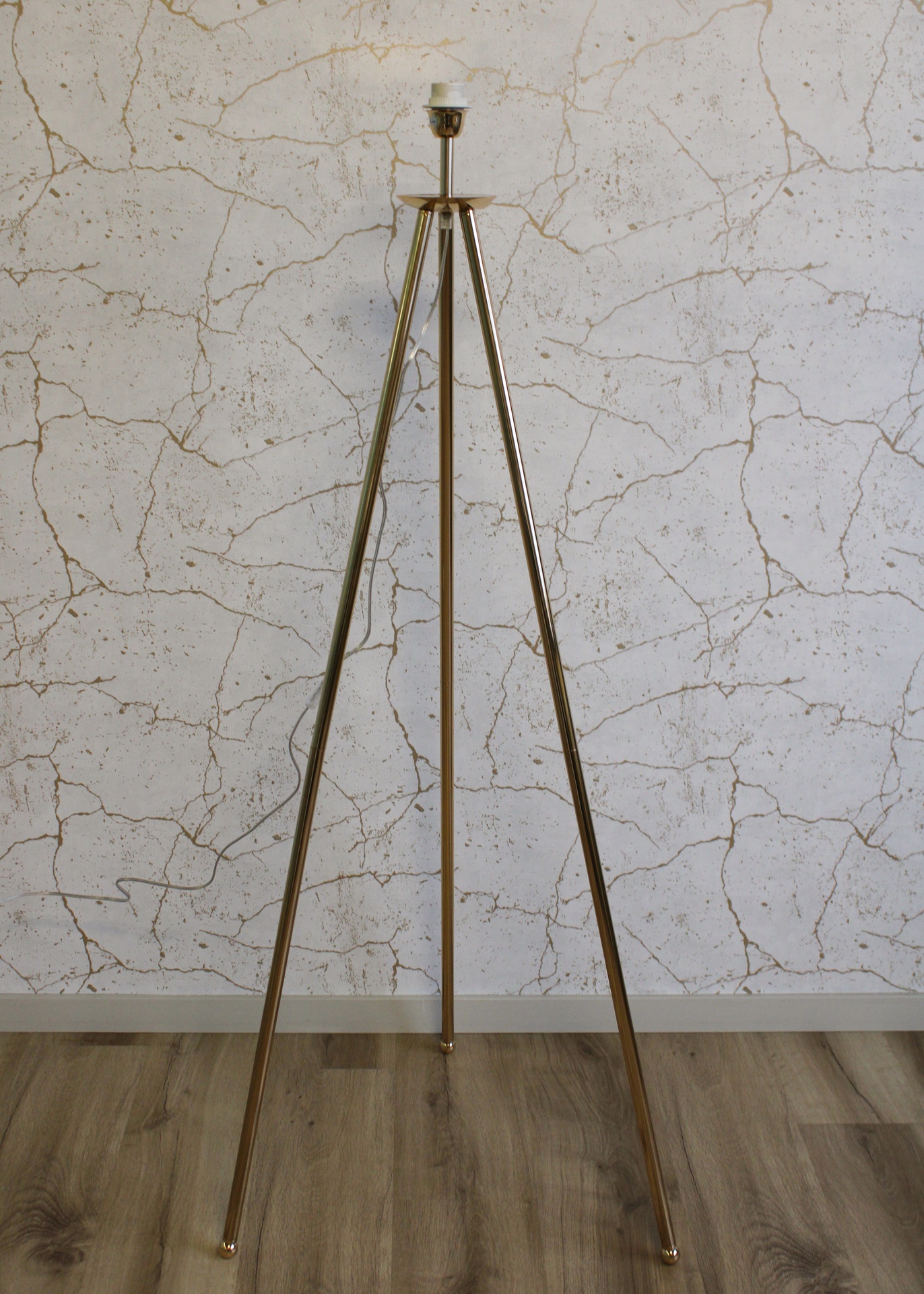 Candeeiro de pé metálico dourado com três pernas e suporte para lâmpada num chão em madeira e parede branca com padrão rachado