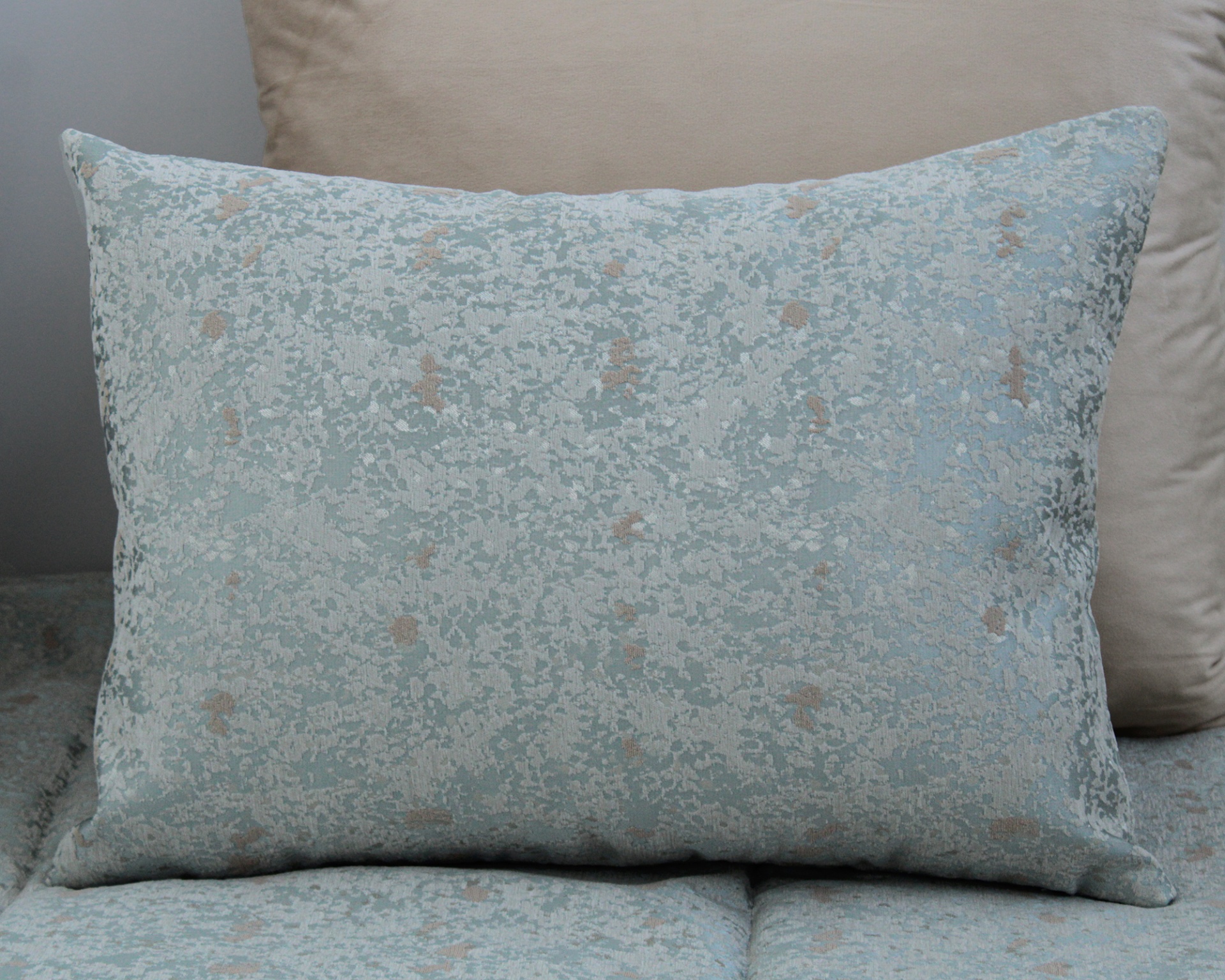 Almofada decorativa retangular azul claro com padrão bege sobre sofá azul com almofada bege.