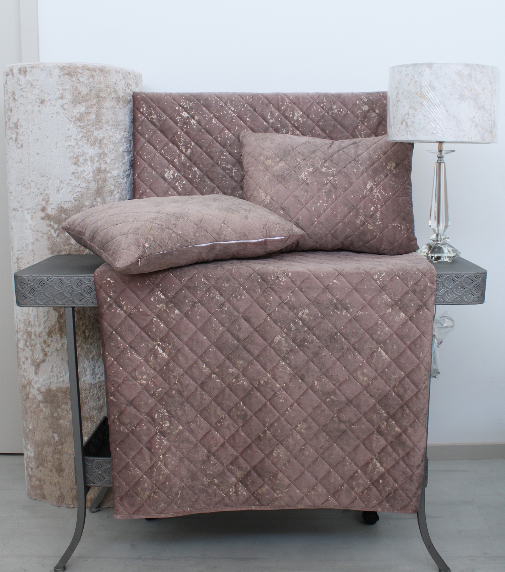 Colcha castanha acolchoada com almofadas, cortinas bege enrolladas, mesa cinza e luminária com abajur branco