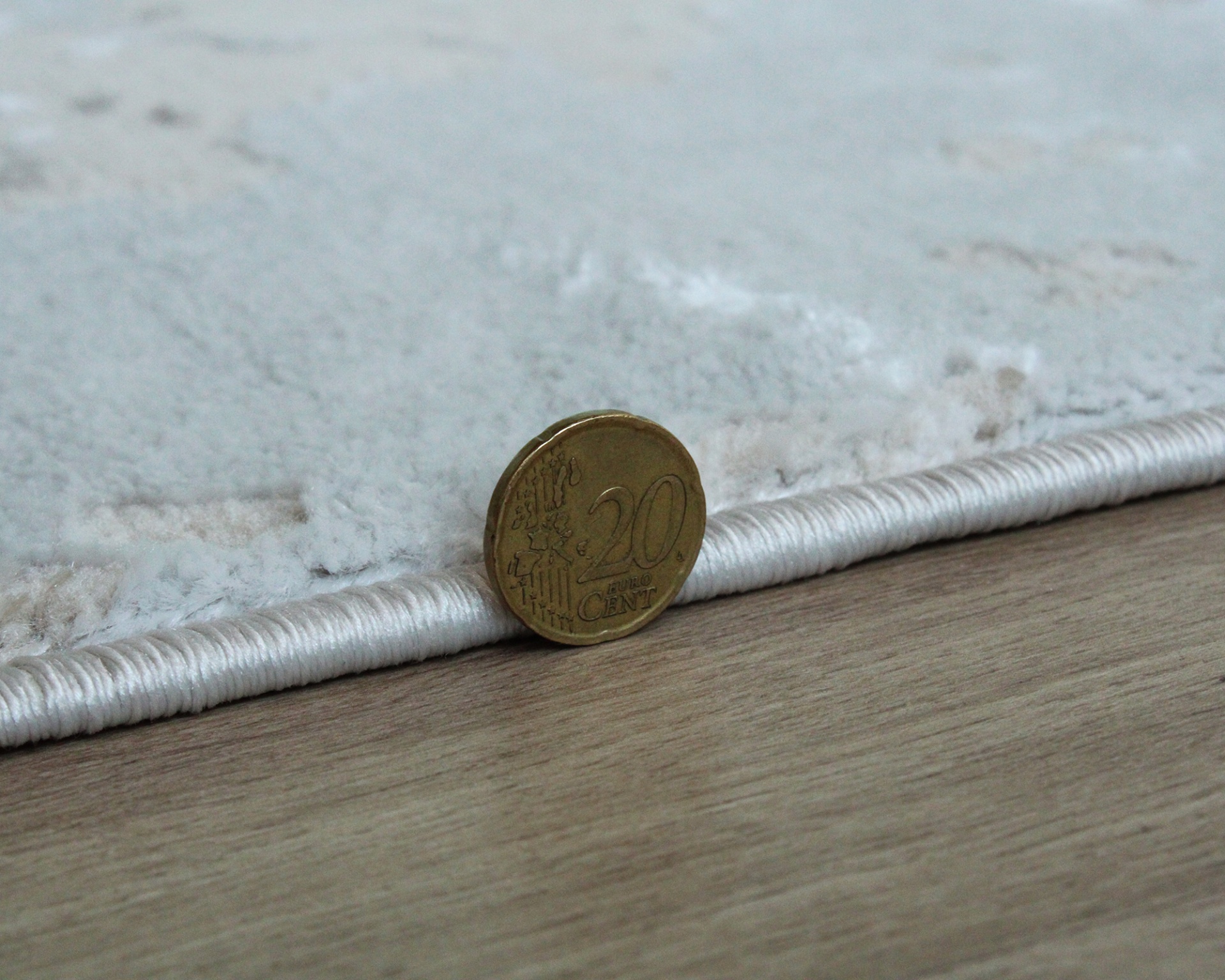 Moeda de 20 cêntimos encostada na borda de tapete branco sobre chão madeira