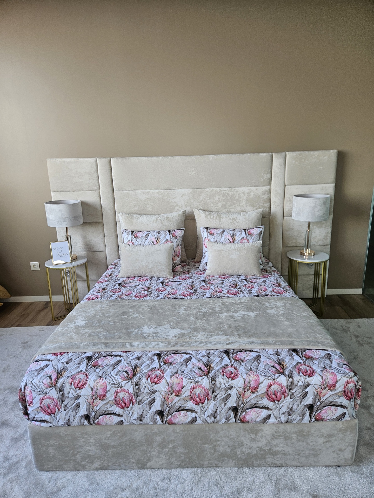 Cama estofada bege com edredão floral, mesas de cabeceira com candeeiros e placa Colibry