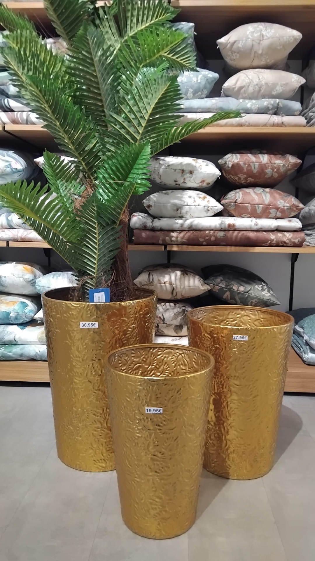 Três vasos dourados em metal com relevo, preços visíveis, um com planta artificial verde, almofadas florais em prateleiras ao fundo.
