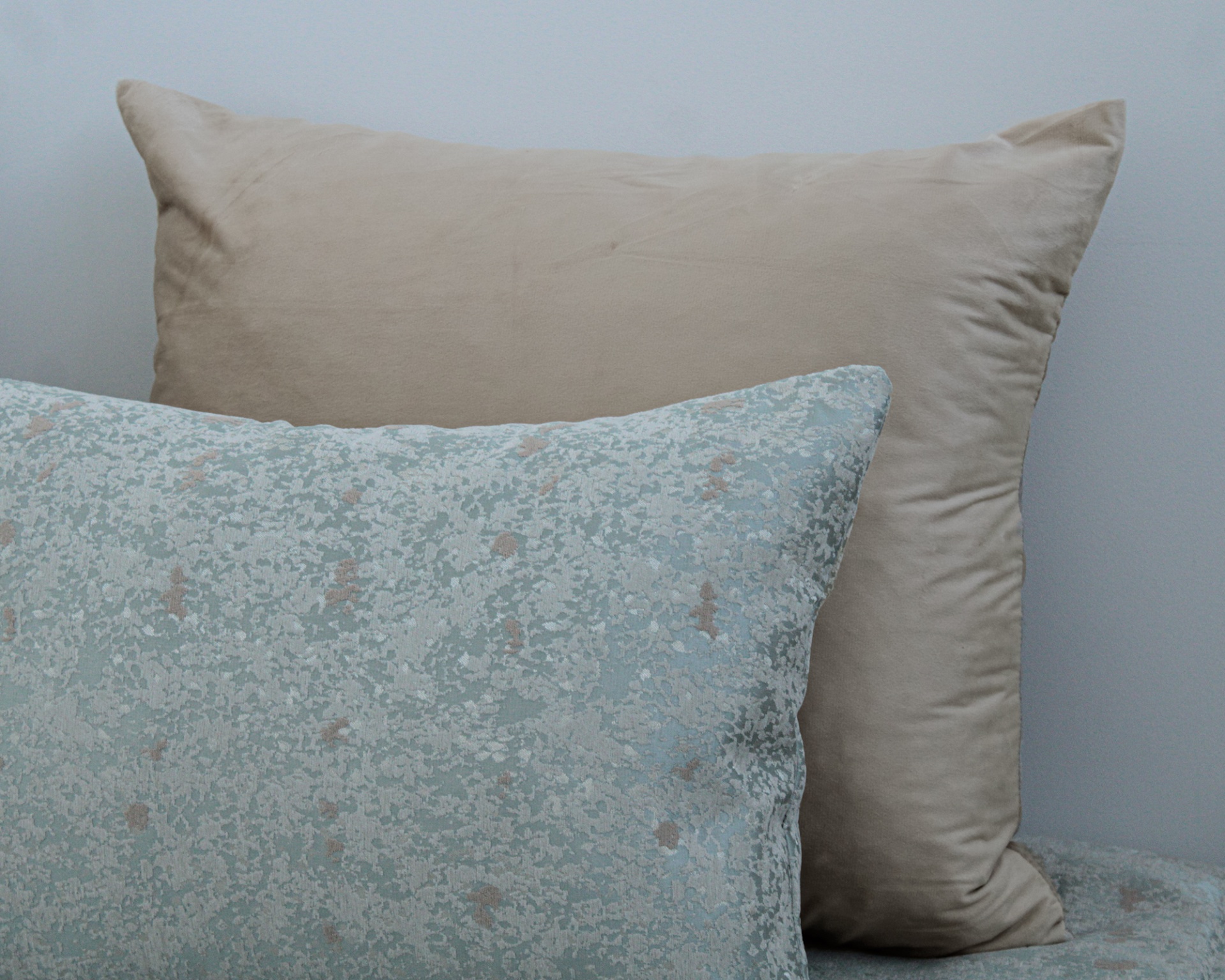 Duas almofadas, uma padrão azul claro com branco e bege e outra lisa bege de veludo.