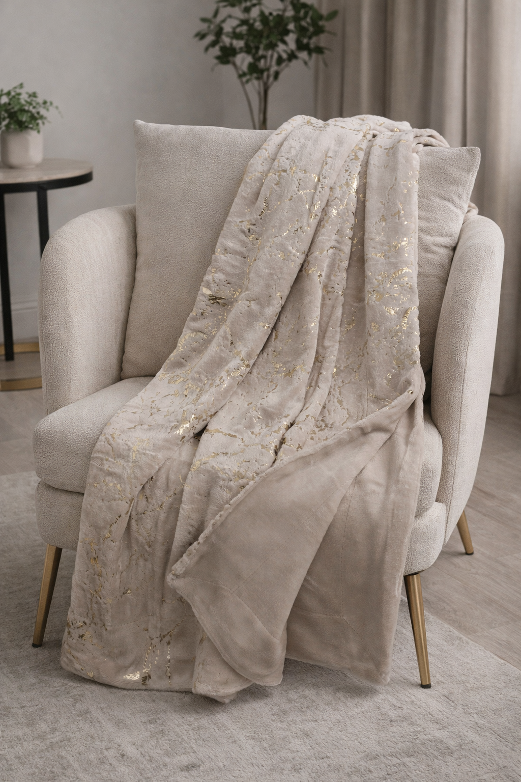 Cobertor bege com padrão dourado sobre poltrona bege em ambiente com plantas.