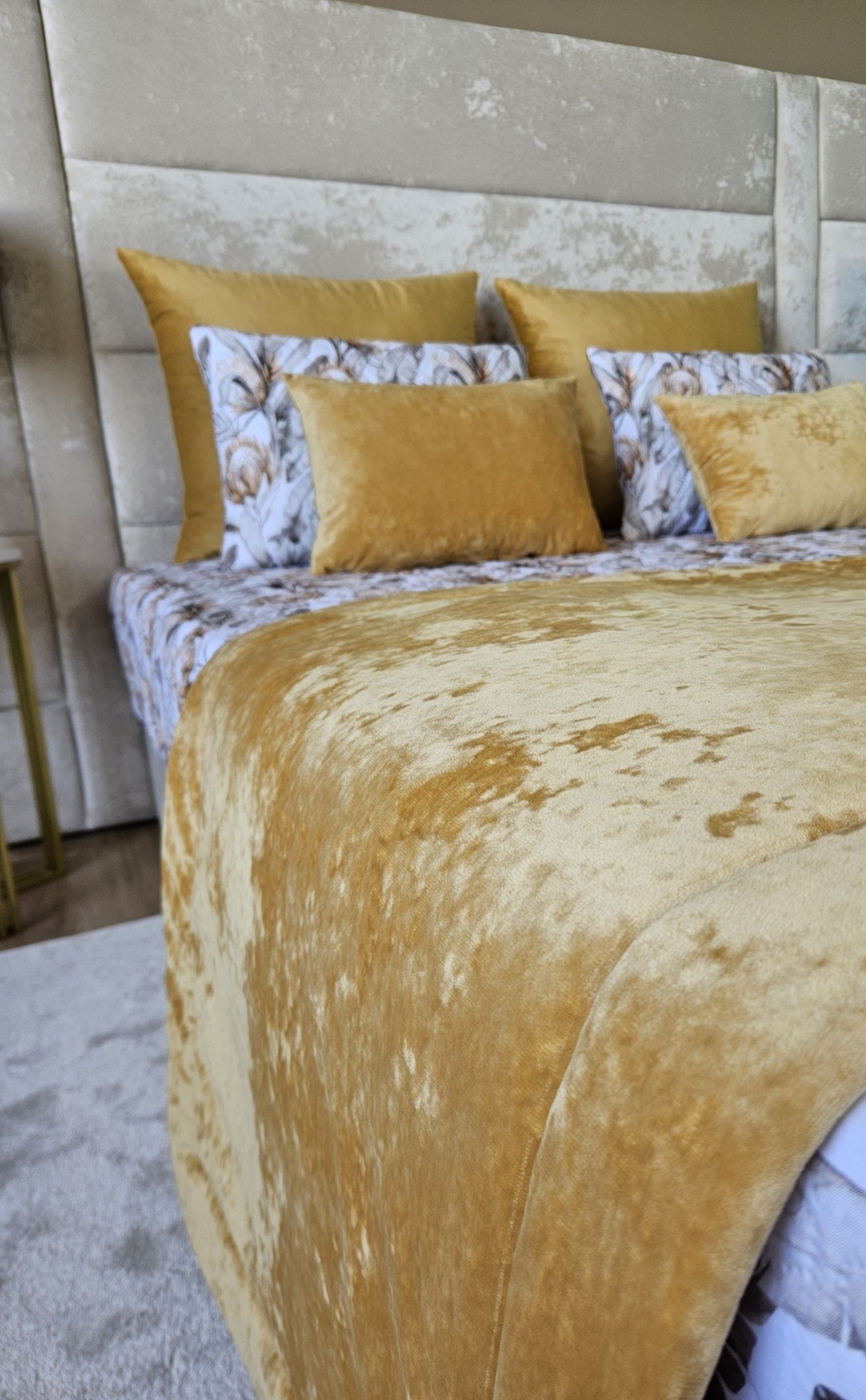 Cama com colcha e almofadas douradas e floridas com cabeceira cinza clara.