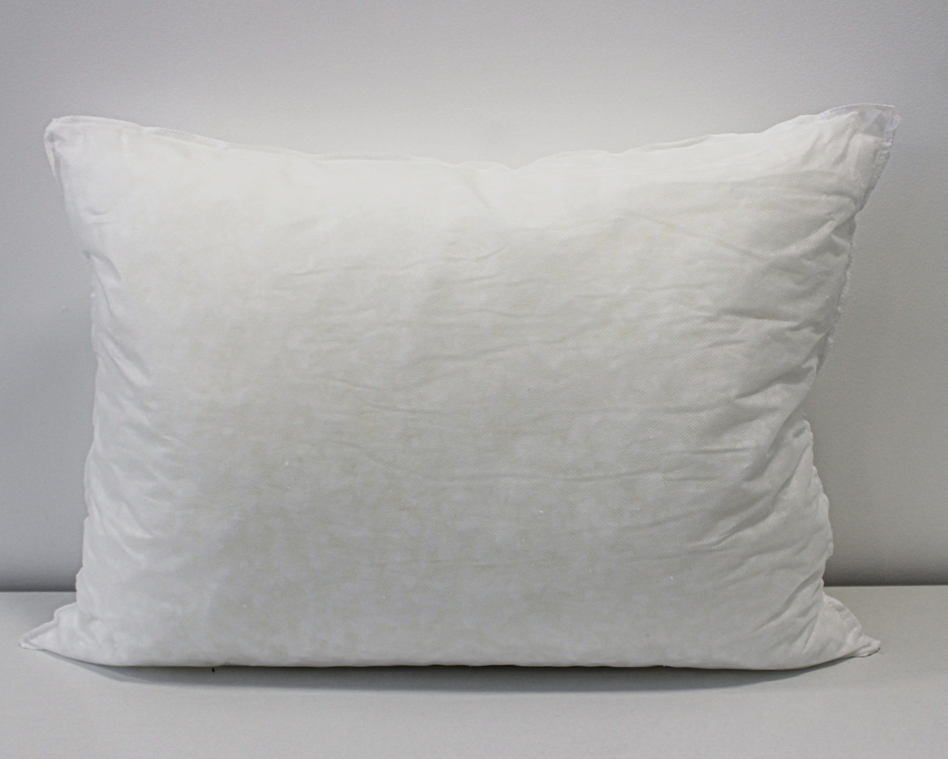 Almofada branca retangular sobre superfície branca e parede cinzenta.