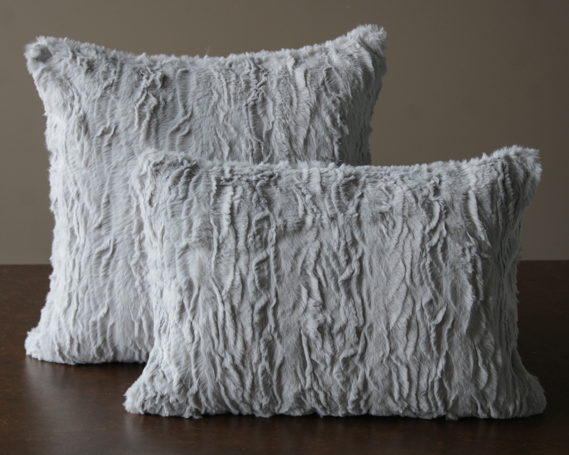 Duas almofadas cinza claras felpudas com textura ondulada sobre mesa de madeira.