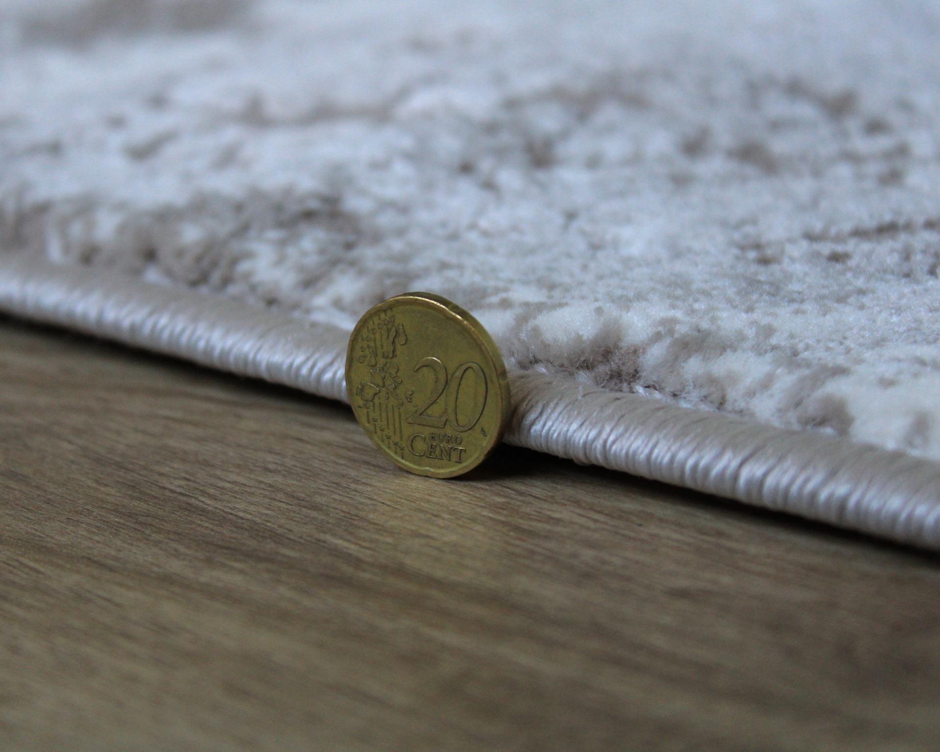 Moeda de 20 cêntimos ao lado da borda de um tapete felpudo claro sobre chão de madeira.