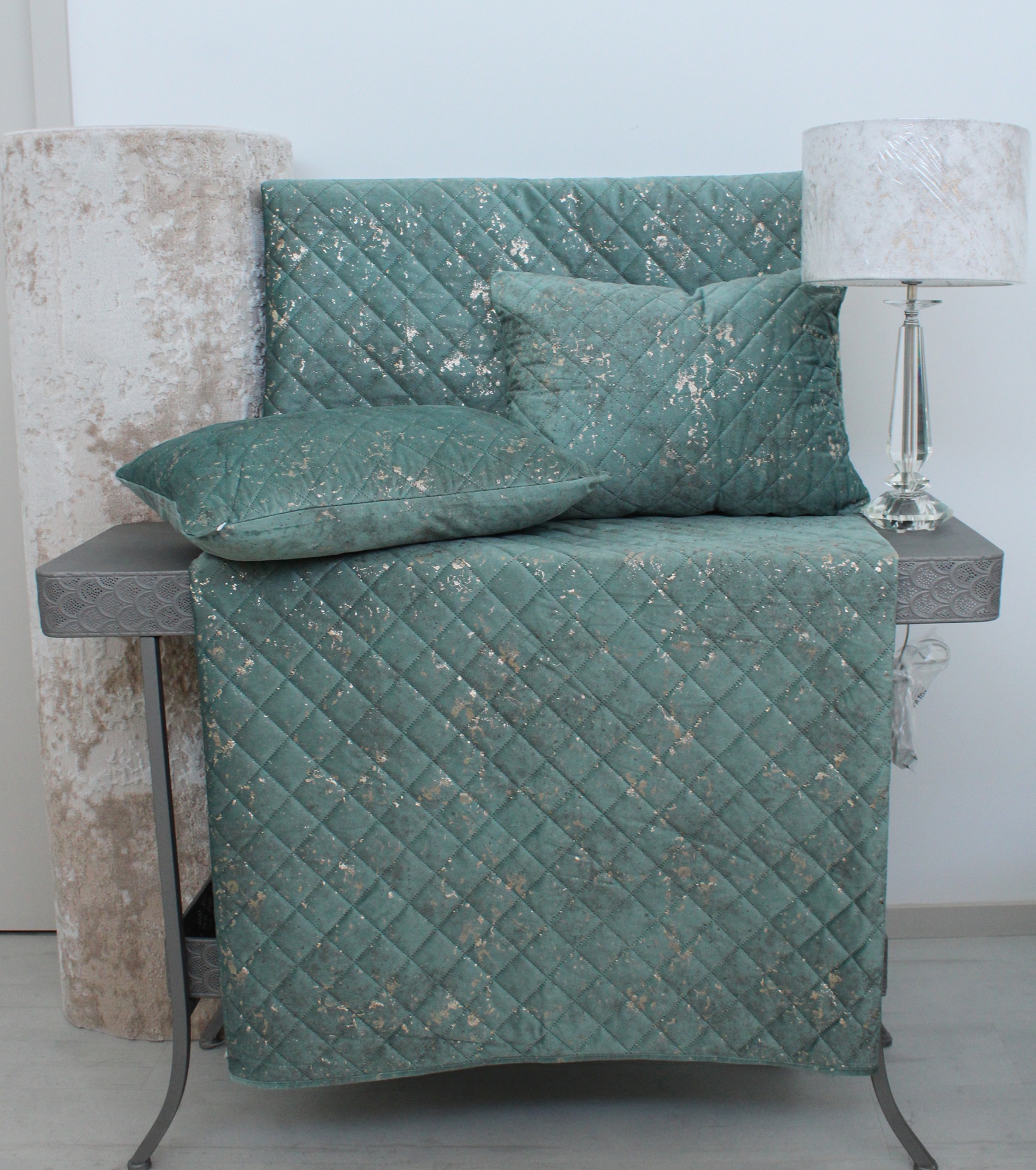 Conjunto de capa acolchoada e almofadas verde-azulado com padrão de diamante e manchas prateadas sobre móvel cinzento e candeeiro com abajur branco.