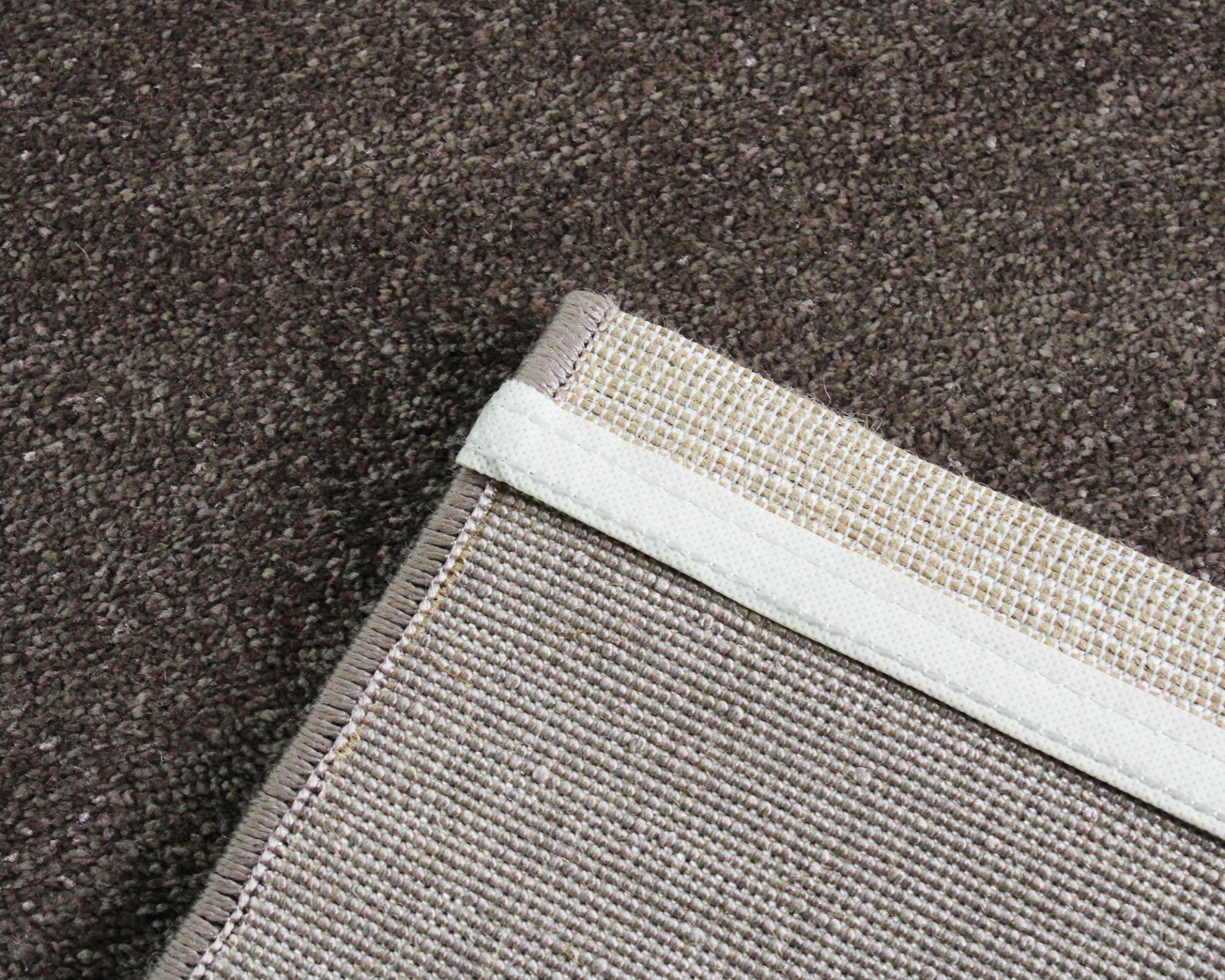 Canto de tapete castanho claro com borda bege sobre carpete castanho escuro.