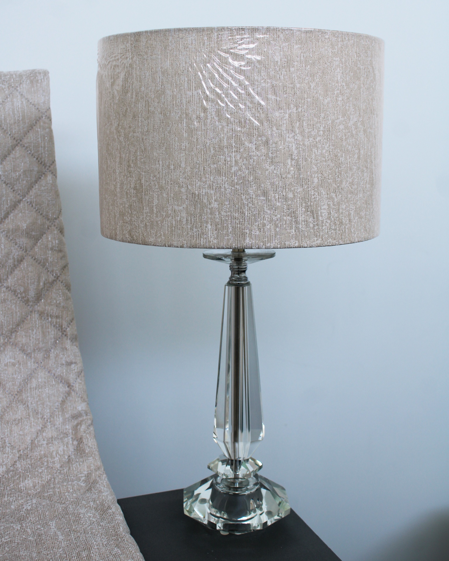 Candeeiro de mesa em cristal com abat-jour bege numa mesa preta ao lado de uma cadeira cinzenta