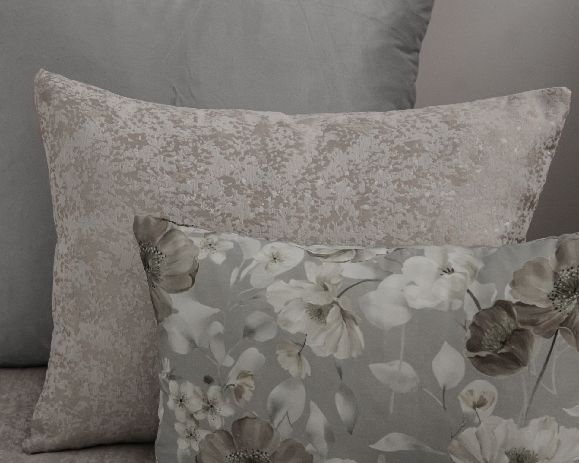 Três almofadas decorativas em tons cinzentos e bege com padrões florais e texturas diferentes