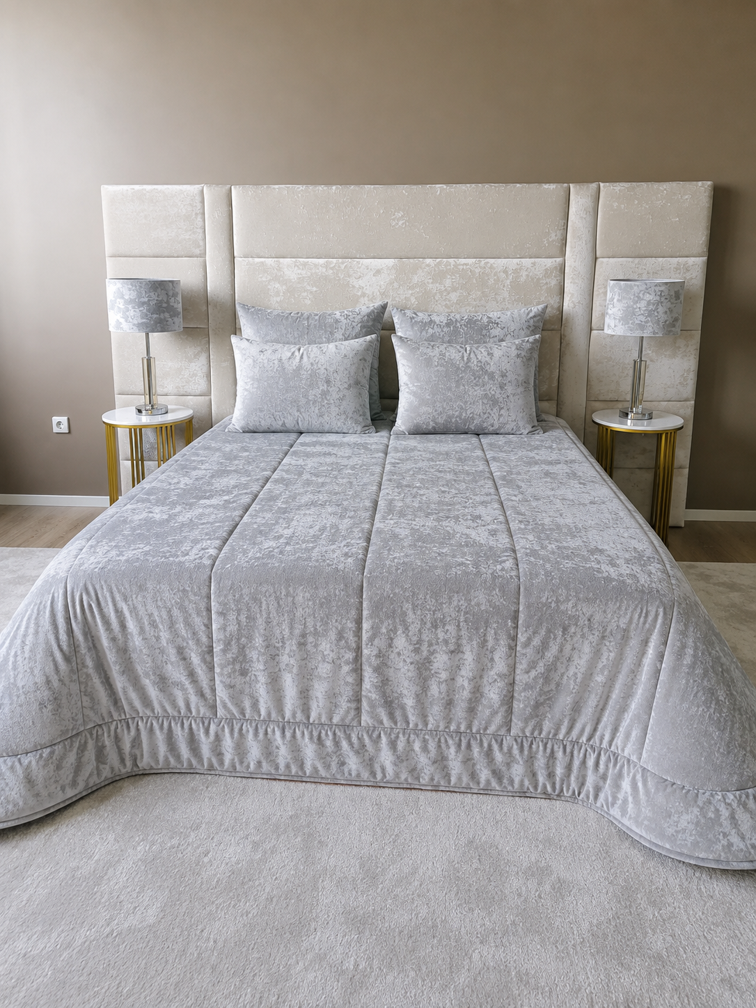 Edredão em Veludo Ref: Paris Cor: Cinza Quarto com cama de casal estofada bege, edredão cinza claro e duas lâmpadas com abajur cinza em mesas laterais brancas.