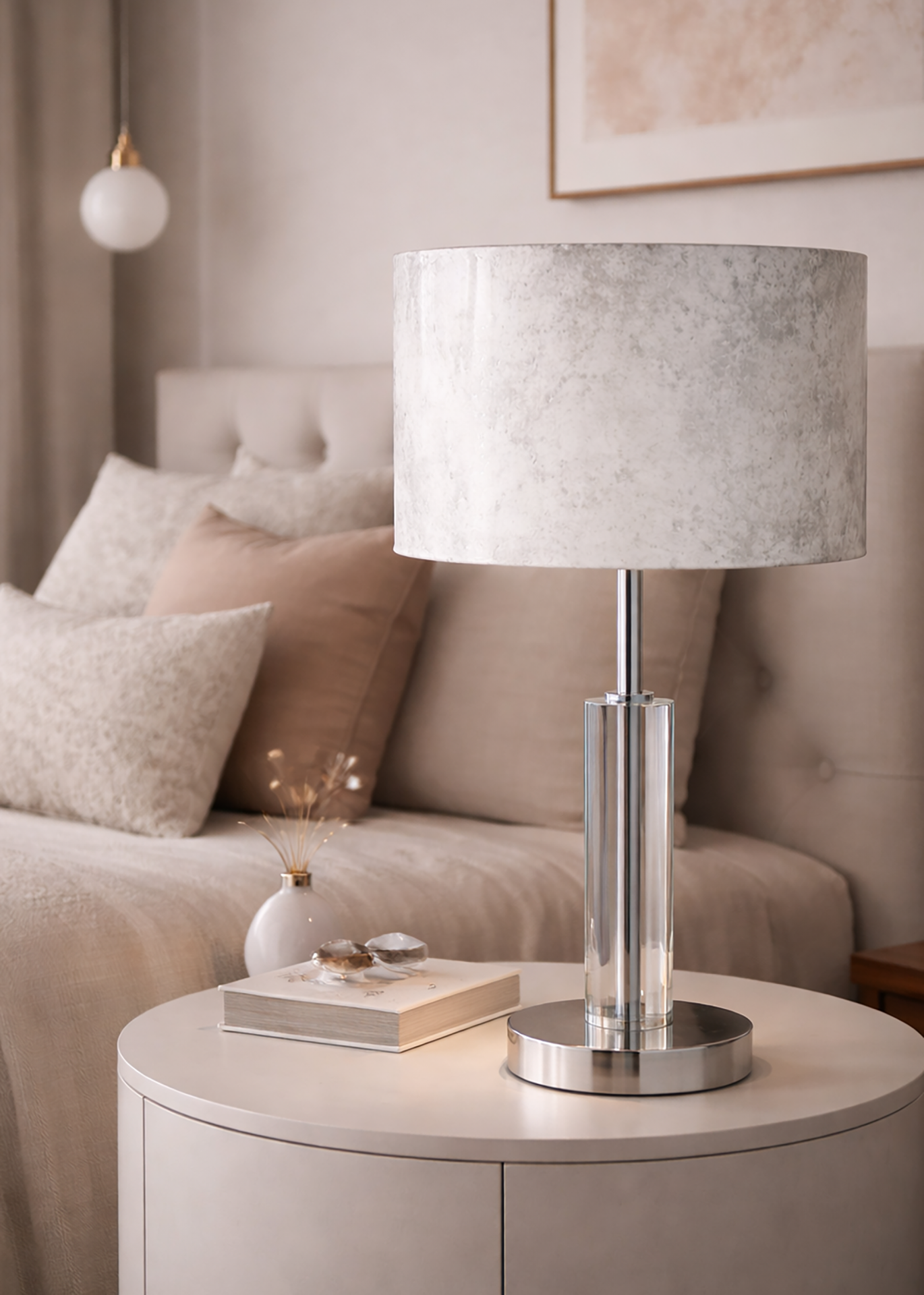 Candeeiro de mesa com base transparente e abat-jour cinza claro numa mesa branca junto a cama