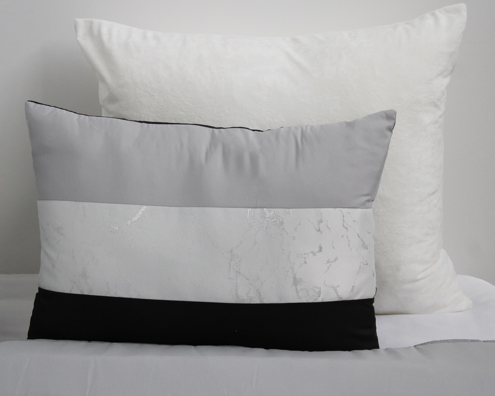 Almofadas em cama, branca atrás e cinza, branco marmoreado e preta à frente.