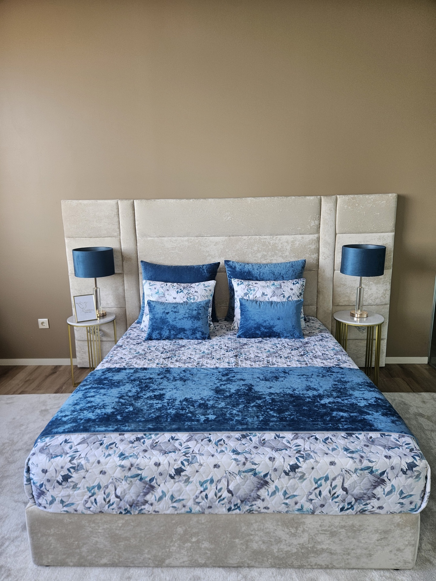 Cama com cabeceira estofada bege, roupa de cama floral e almofadas azuis, mesas laterais douradas, candeeiros azuis, parede castanha.