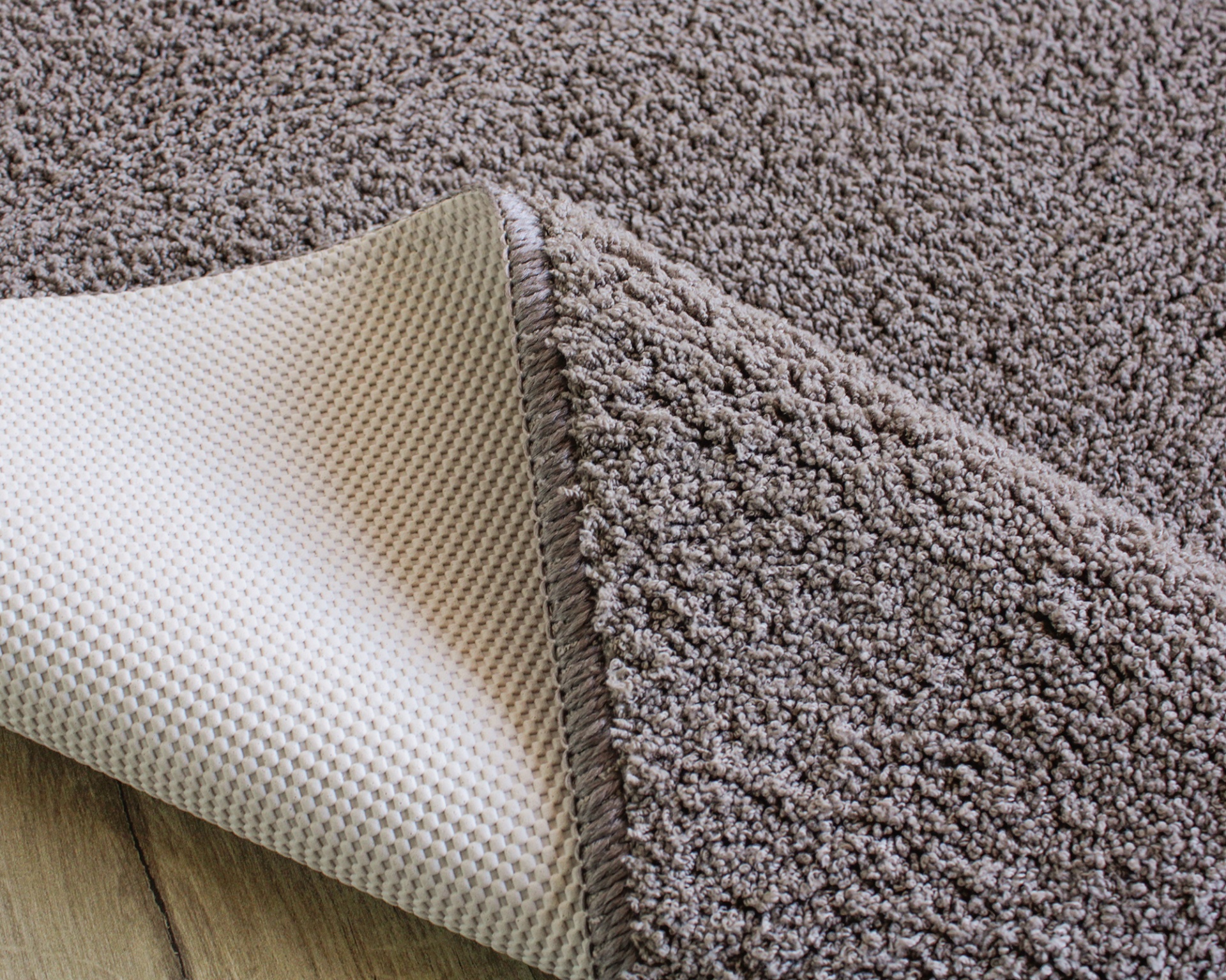 Tapete castanho claro felpudo com base antiderrapante bege sobre piso de madeira.