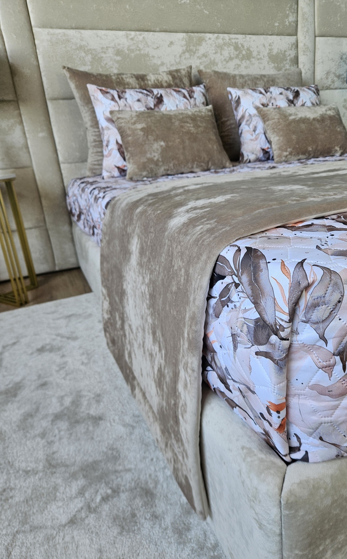 Cama estofada bege com almofadas castanhas e padrão de folhas, edredão castanho claro e tapete bege.