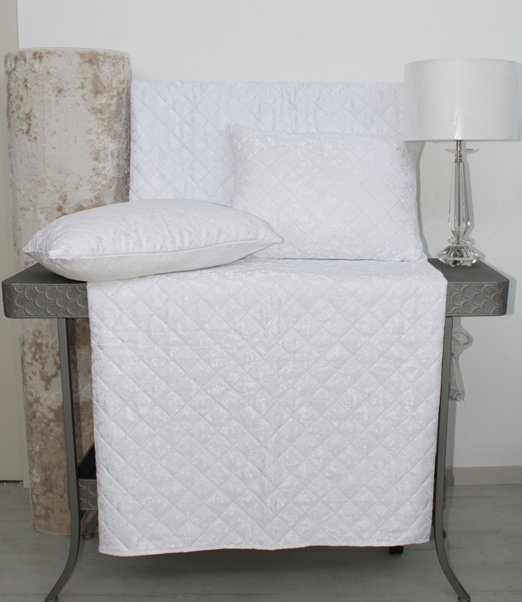 Conjunto de cama branco acolchoado com padrão em losango sobre mesa cinza ao lado de candeeiro de mesa