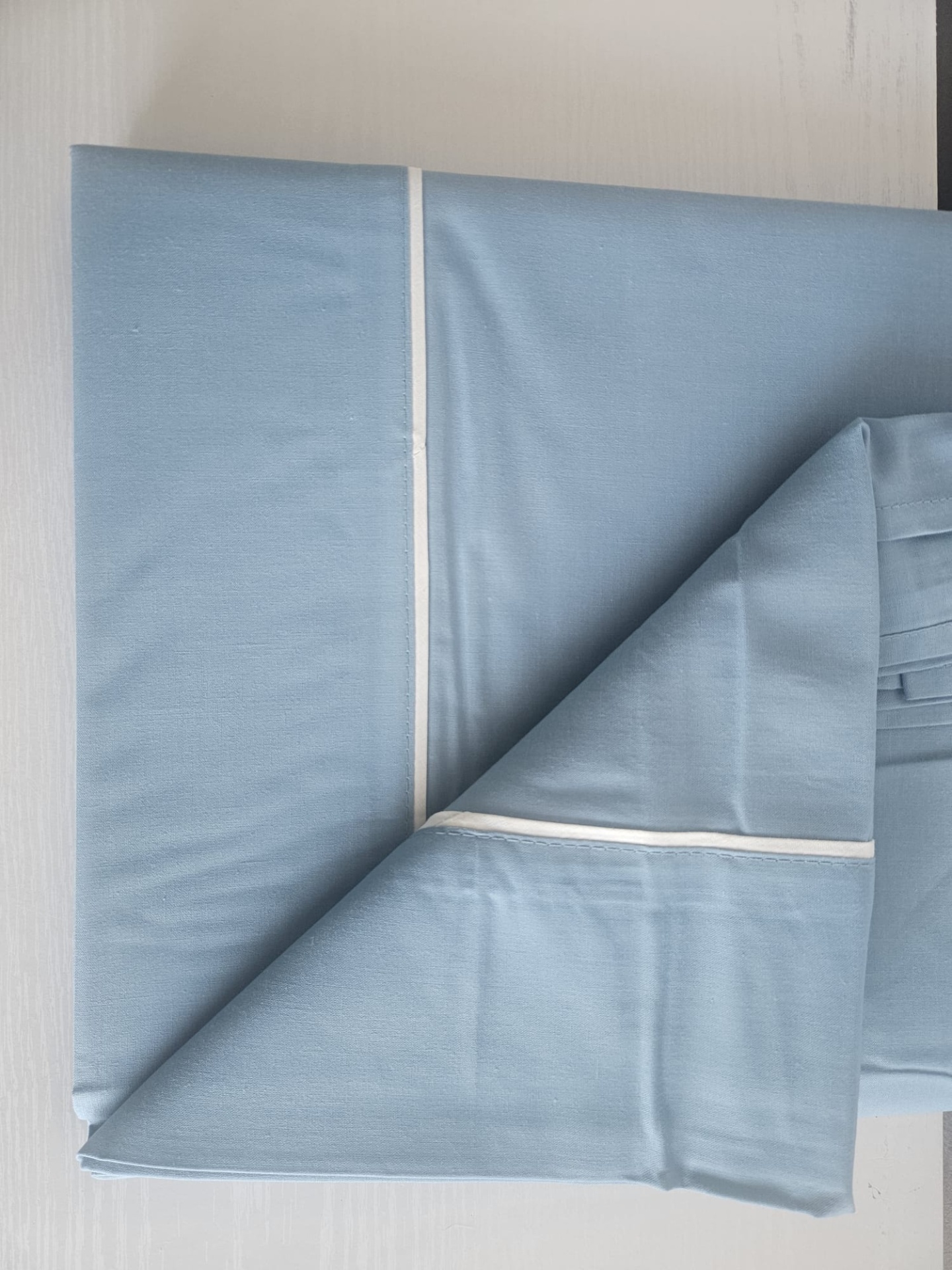 Roupas de cama azul claro com linha branca dobradas em superfície clara