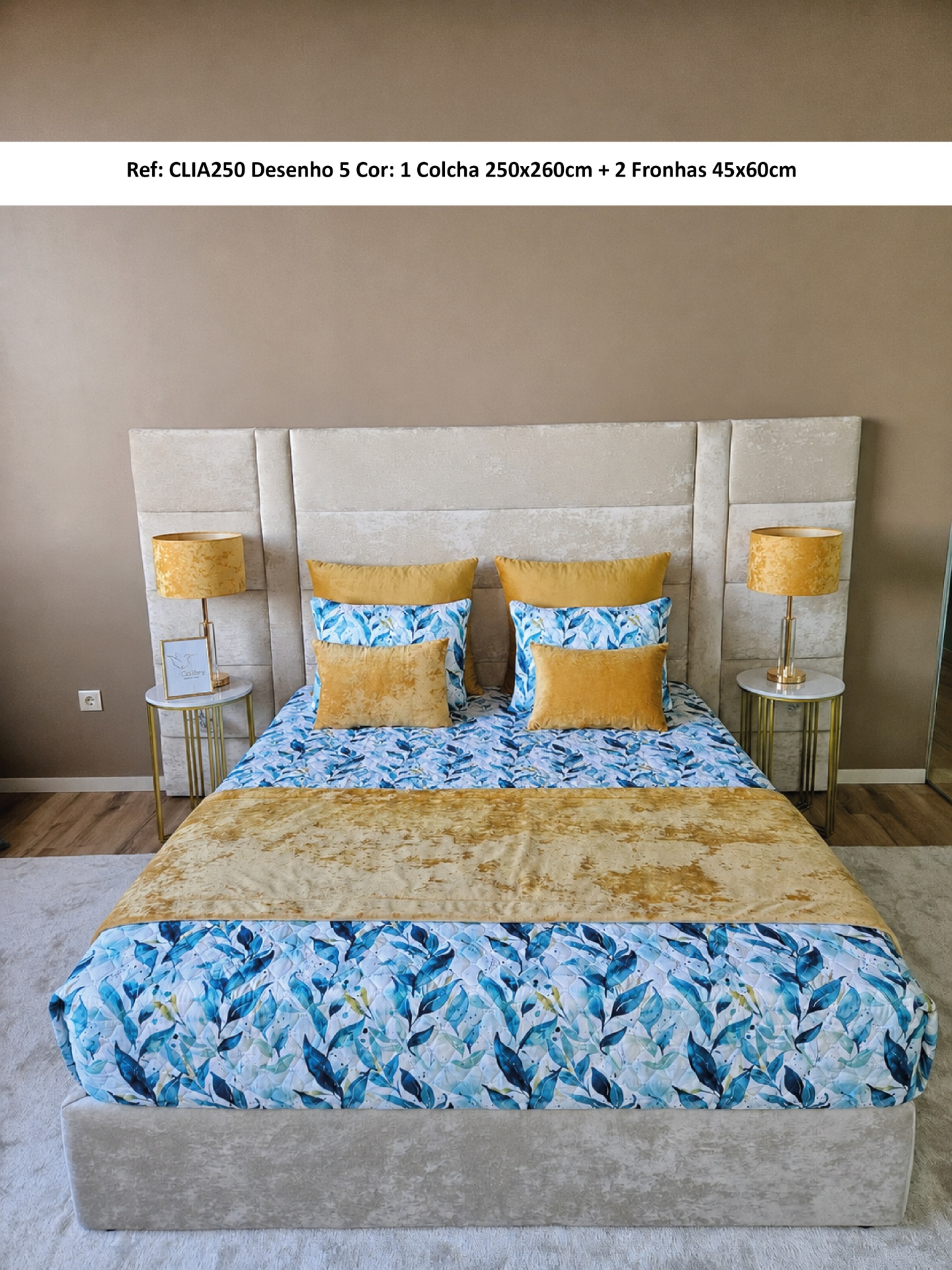 Cama com colcha floral azul e verde e almofadas amarelas, cabeceira estofada bege, mesas e candeeiros laterais amarelos.