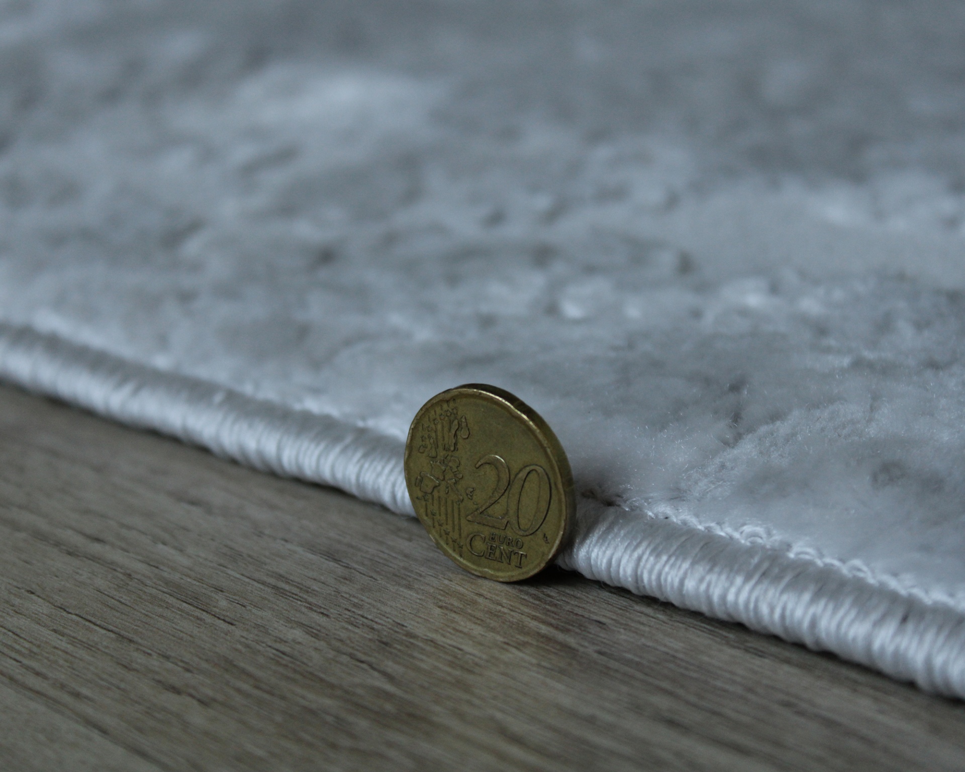 Moeda de 20 cêntimos ao lado de tapete cinzento claro sobre chão de madeira