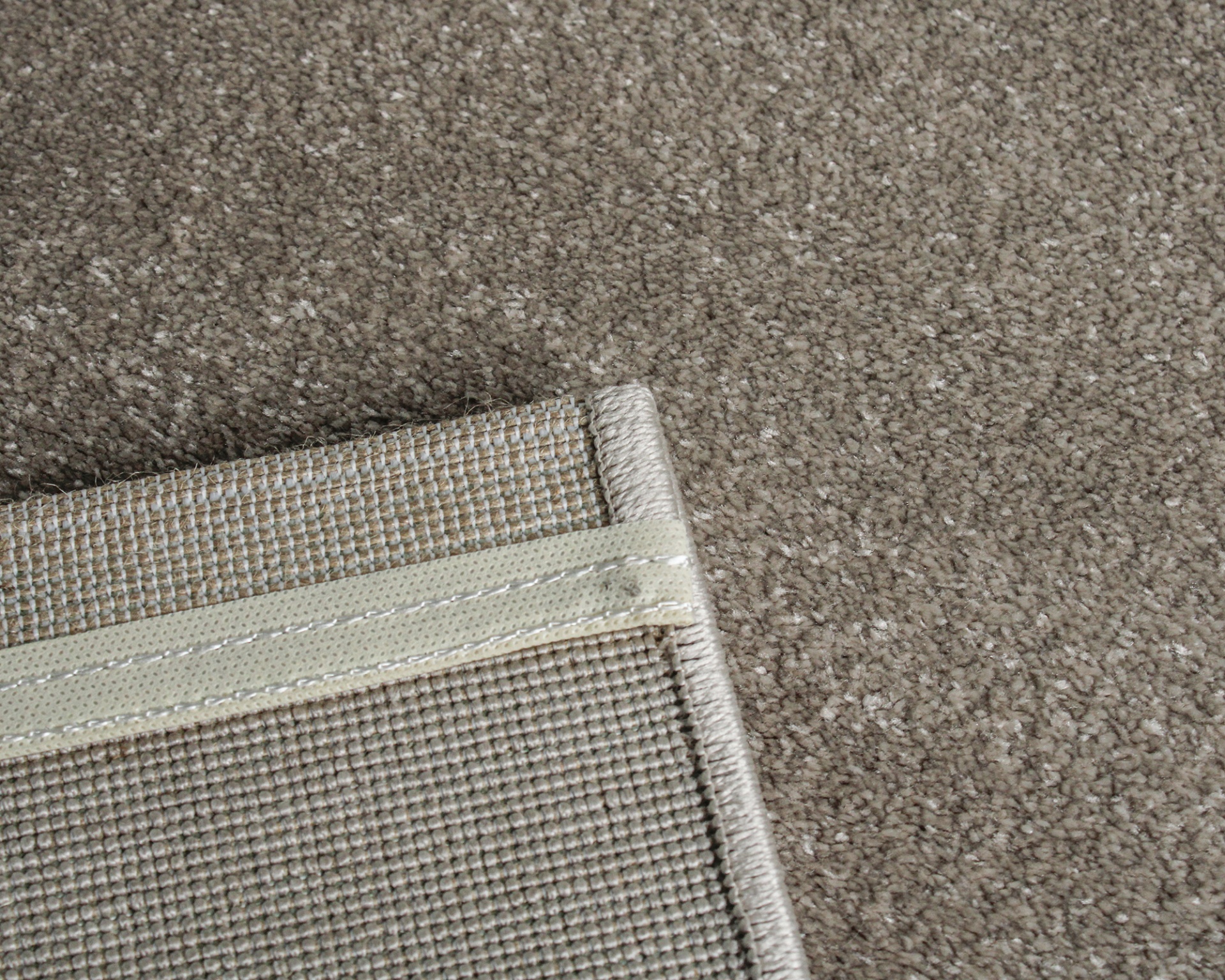 Tapete cinzento claro com verso tecido bege e branco, bordas costuradas.