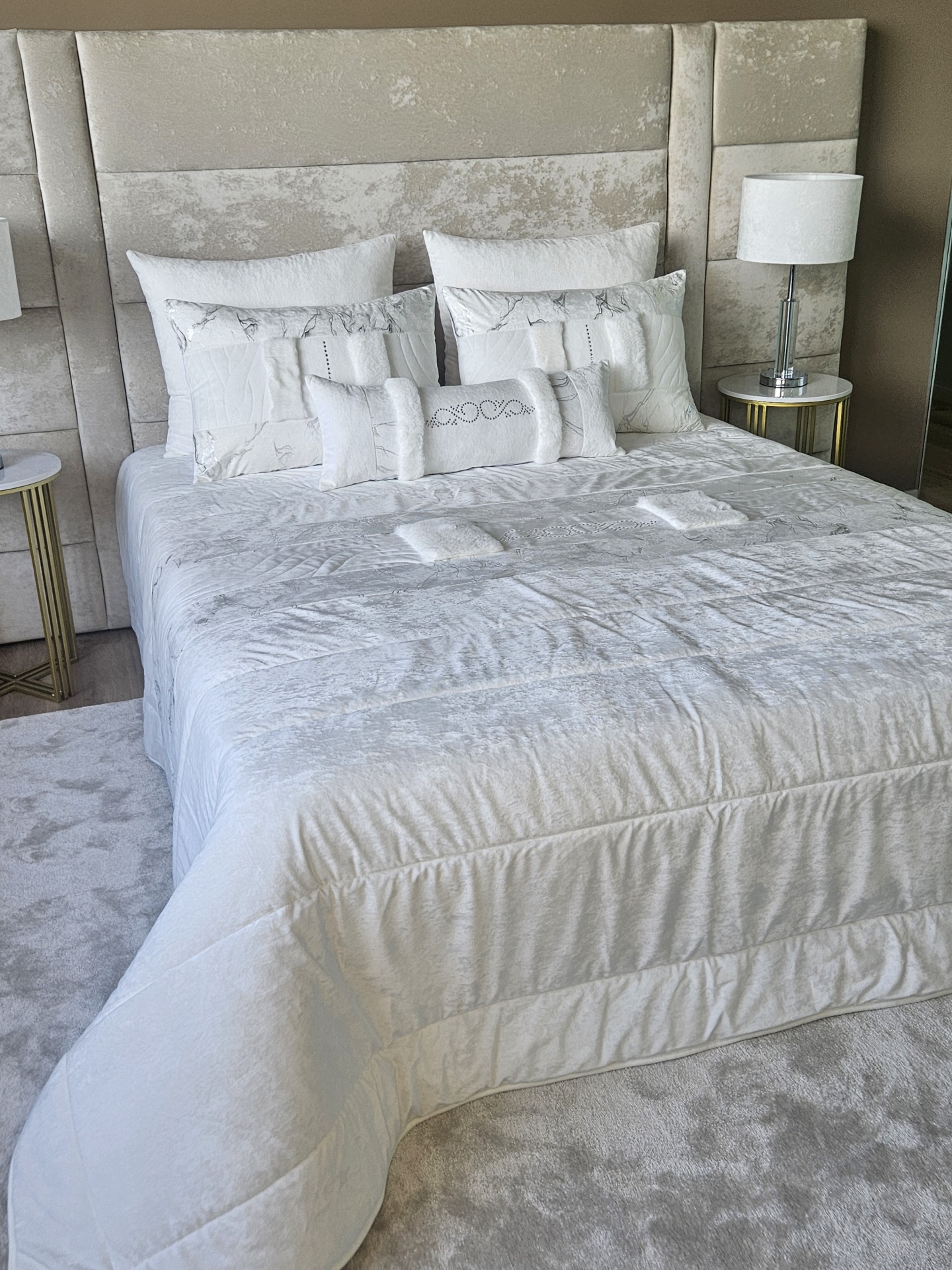 Edredão em Veludo Ref: Nepal Branco Cama com conjunto branco e cabeceira estofada cinza claro, almofadas brancas e mesas de apoio com candeeiros.