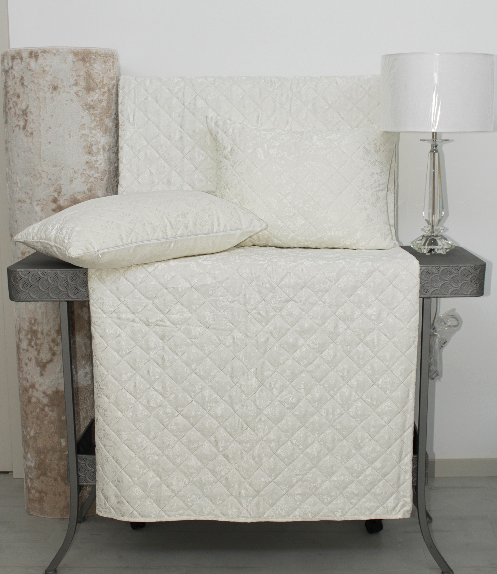 Cobertura e almofada acolchoadas em tecido acetinado branco com padrão de losangos sobre mesa escura, com rolo de tecido bege e candeeiro branco ao lado