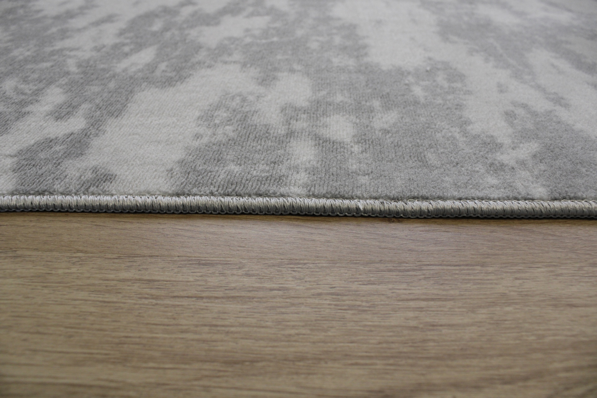 Tapete cinzento claro com padrão abstrato e bordas costuradas sobre chão de madeira