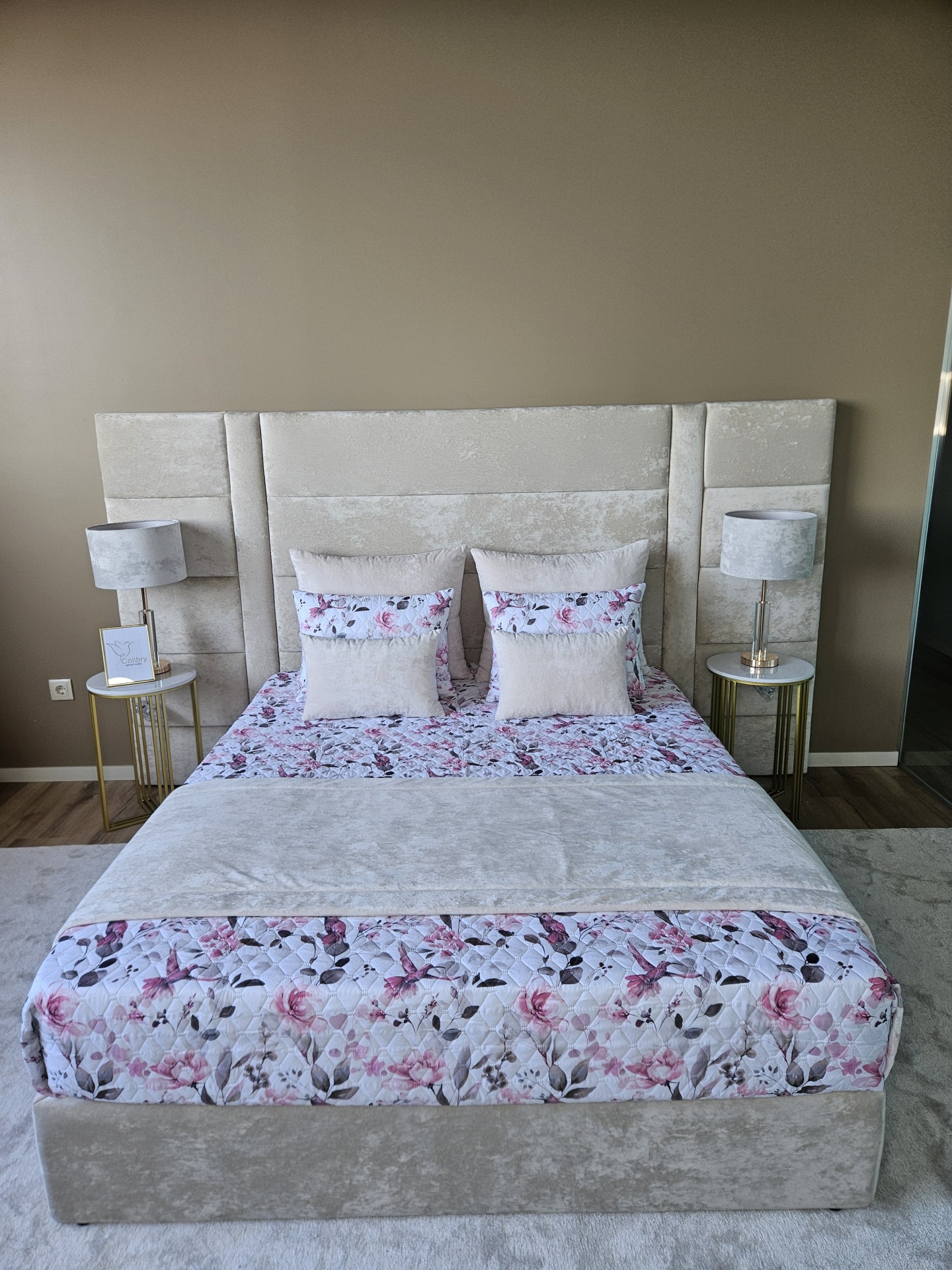 Cama estofada creme com roupa de cama floral em rosa e lilás e duas mesas de cabeceira com lâmpadas cinza