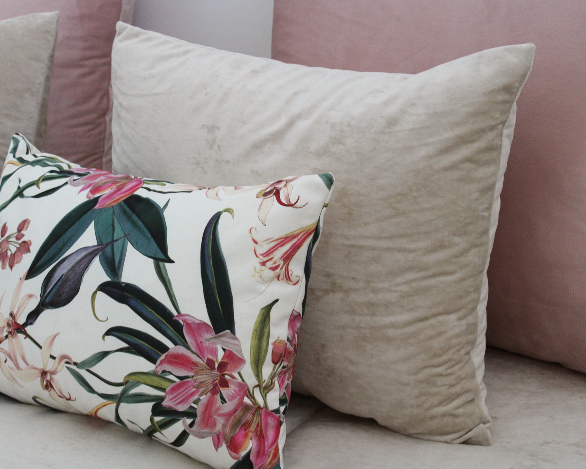 Três almofadas decorativas com padrão floral, bege aveludado e rosa aveludado sobre sofá claro.