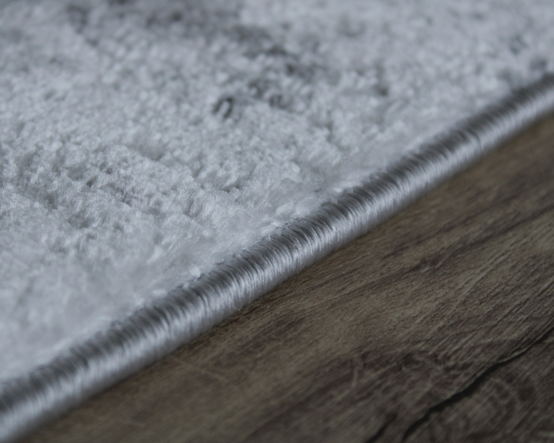 Tapete cinza claro com textura macia, borda enrolada, sobre chão de madeira.
