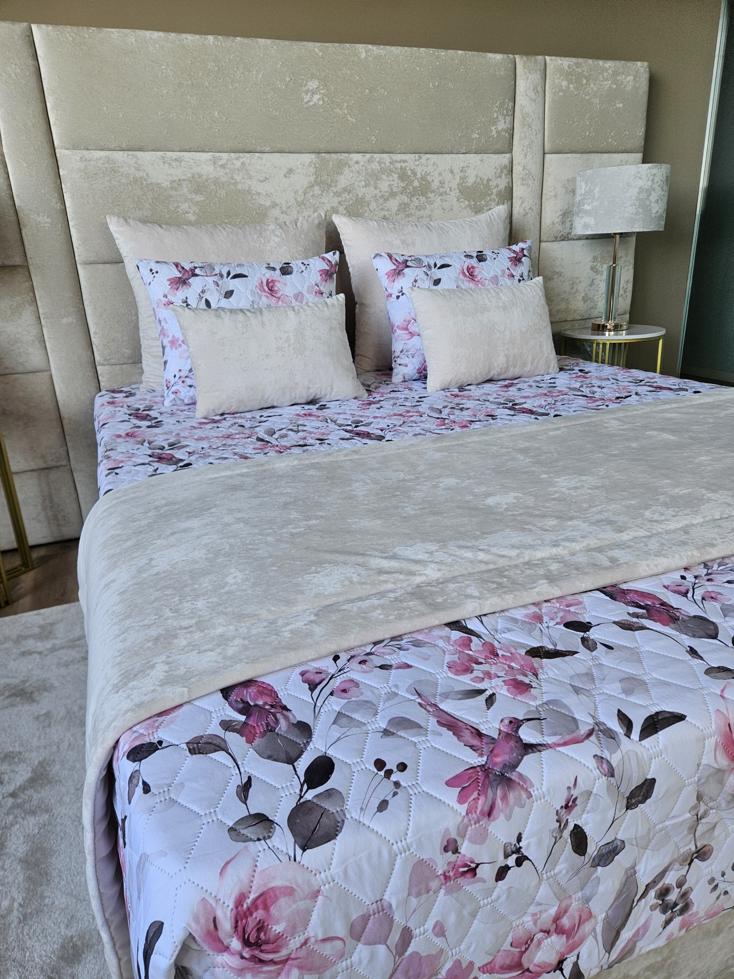 Cama com roupa de cama floral e almofadas beije com cabeceira estofada e candeeiro ao lado.