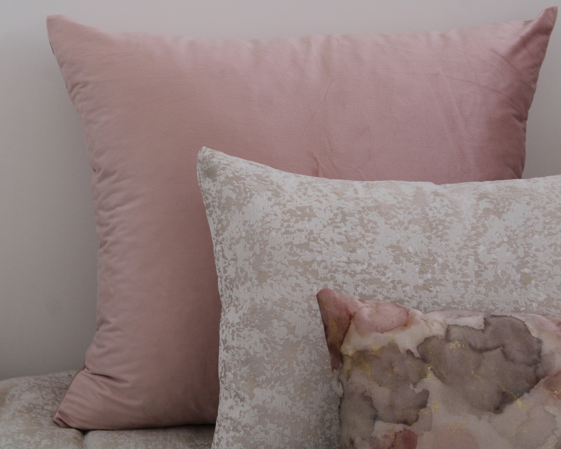 Três almofadas decorativas em tons rosa, bege e cinza com diferentes texturas e padrões