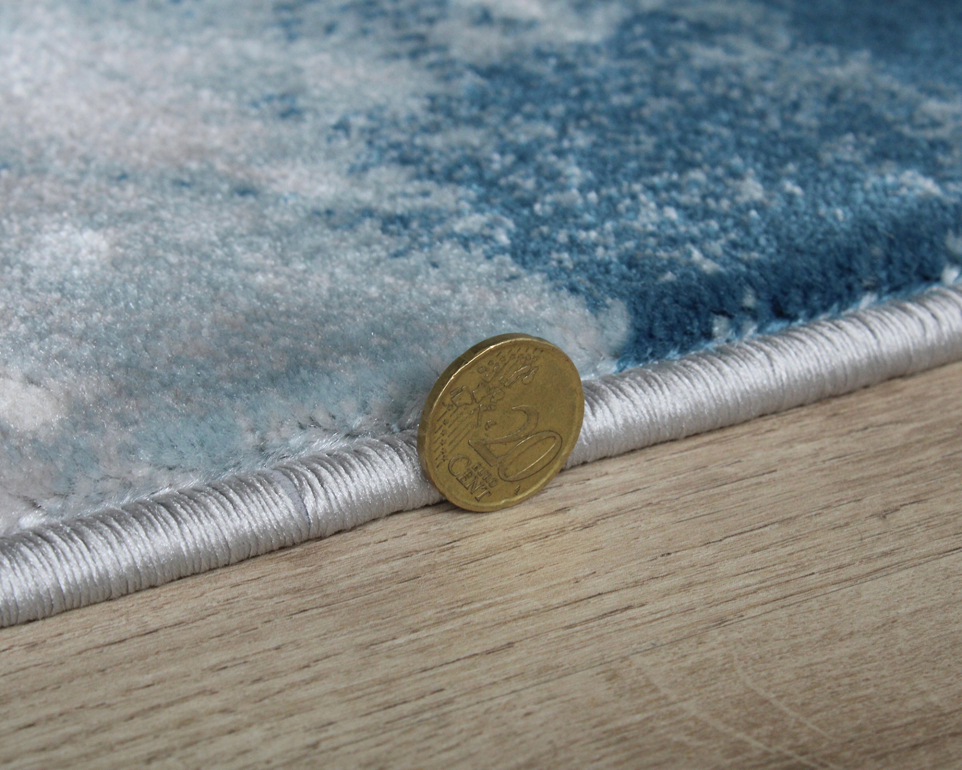 Tapete azul e branco com borda em foco, moeda de 20 cêntimos ao lado no chão de madeira