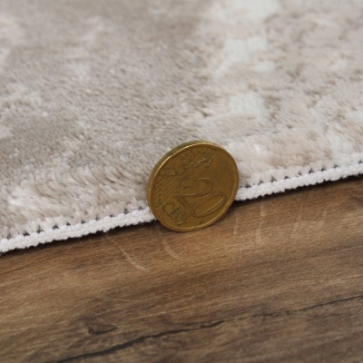 Moeda de 20 cêntimos junto a tapete bege com padrão floral em relevo sobre chão de madeira.