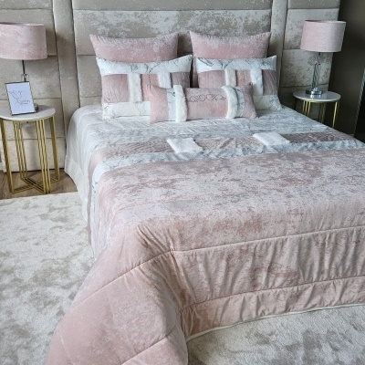 Cama com roupa de cama rosa e bege aveludada e almofadas decorativas com padrão em retângulos, cabeceira estofada bege, mesas laterais douradas com tampo branco e candeeiros rosa.