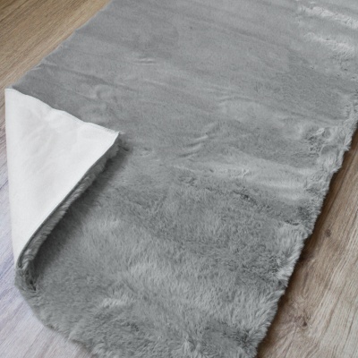 Tapete cinzento peludo com canto dobrado, sobre chão de madeira clara