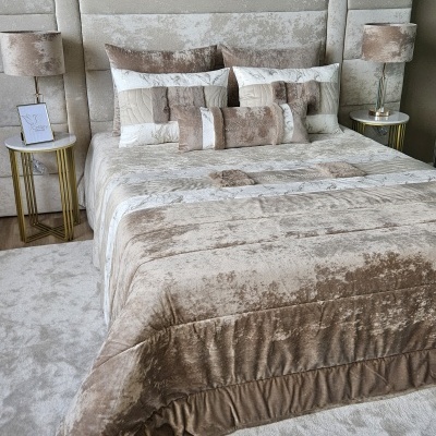 Cama estofada em veludo com roupa e almofadas em tons bege e creme, mesas de cabeceira douradas com candeeiros.
