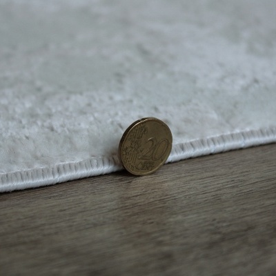Tapete branco felpudo com moeda de 20 cêntimos encostada para comparação de espessura sobre chão de madeira.