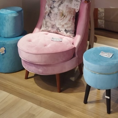 Cadeira rosa de veludo com almofada e duas pufes redondas azuis com etiquetas, tudo sobre chão de madeira.