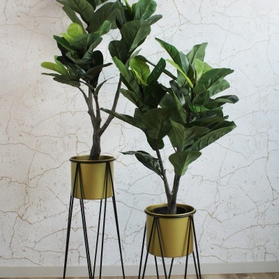 Dois vasos dourados com suportes pretos e plantas artificiais verdes em chão de madeira